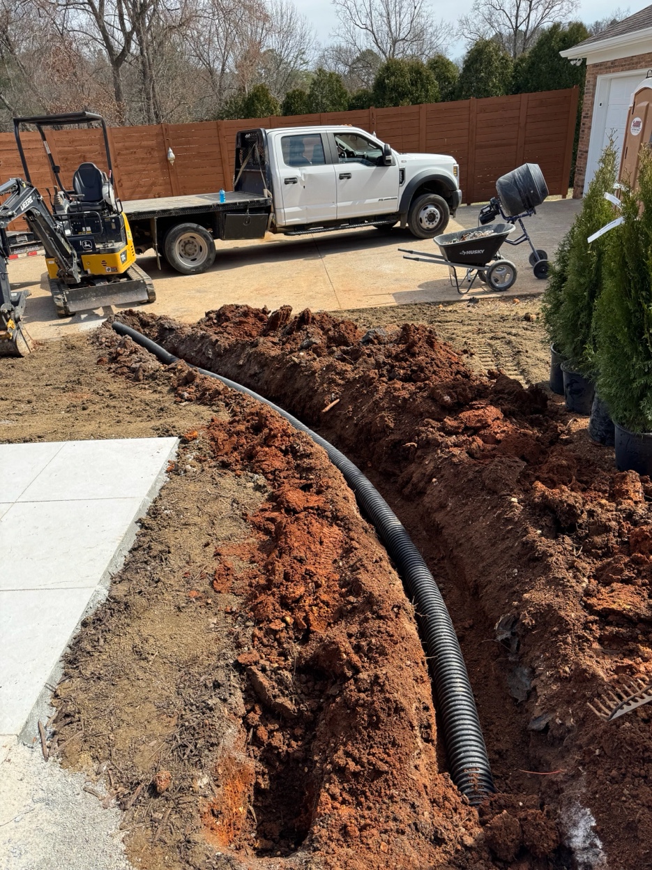 Ray Lawns crew excavating a drainage trench with a mini excavator and work truck on site in Ooltewah Tennessee