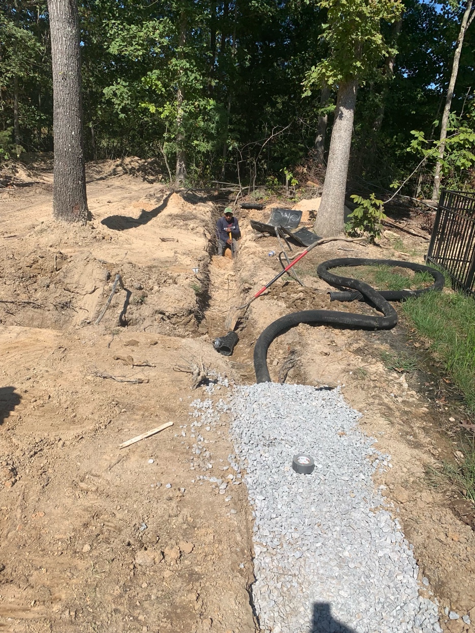 Crew member working in the drainage trench with gravel fill and perforated pipe with filter sock visible during French drain installation